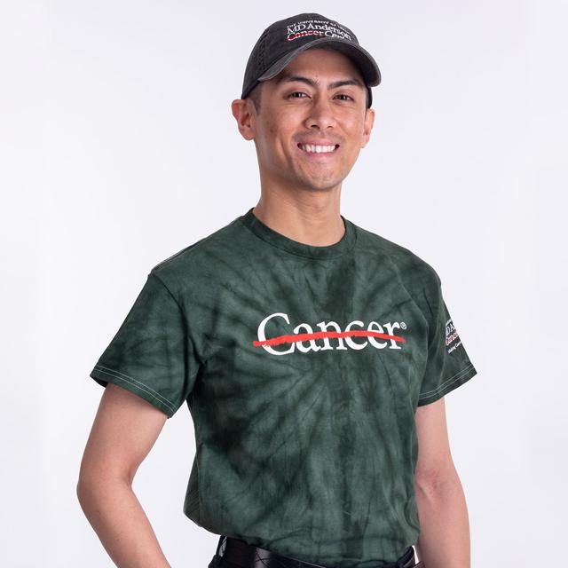 MD Anderson employee wearing a green tie-dye shirt with the white cancer strikethrough logo on the chest and the full MD Anderson logo on the sleeve, paired with a black denim baseball cap featuring the white MD Anderson logo.